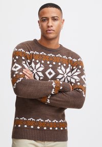 Suéter marrón de punto con patrones geométricos en blanco y naranja, cuello redondo, puños y dobladillo acanalados, que presenta intrincados diseños inspirados en el invierno.