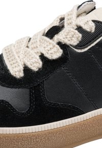 Κοντινή λήψη μαύρου αθλητικού παπουτσιού με χονδρά κρεμ  πλεγμένα κορδόνια και υφή στο ανοιχτό καφέ πέλμα.