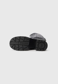 Μαύρη σόλα από καουτσούκ μποτών με άνισο σχέδιο πέλματος, που διαθέτει στρογγυλό σχήμα και ελαφρύ τακούνι. Είναι ορατό το λείο γκρι ανώτερο υλικό.