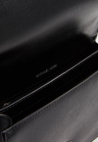 Interno di una borsa a tracolla in pelle nera con tasca con zip e marchio in rilievo "Michael Kors". Texture liscia con linee pulite e design minimalista.
