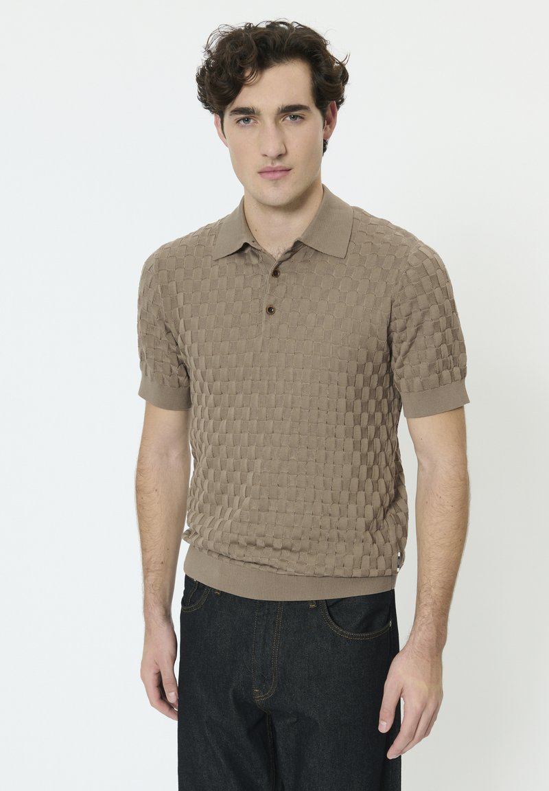 Jeune homme aux cheveux bouclés foncés portant un polo beige texturé à manches courtes et un jean bleu foncé, debout devant un fond blanc.