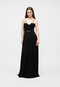 Γυναίκα με μακριά καστανά μαλλιά, φορώντας μακρύ μαύρο πλισέ φόρεμα με λεπτούς ιμάντες και εφαρμοστό κορσέ, στέκεται μπροστά σε απλό φόντο.