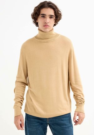 Suéter de cuello alto beige hecho de un material de punto suave, con mangas largas y un ajuste relajado, combinado con jeans azules.