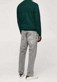 Personne portant un jean gris clair, un sweat vert foncé et des baskets blanches à talons marron, debout devant un fond clair uni.