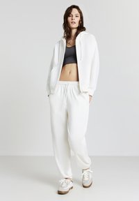 Junge Frau trägt einen weißen Kapuzenpullover, weiße Jogginghose, grauen Sport-BH und weiße Sneakers und steht vor einem schlichten grauen Hintergrund.