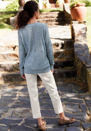 Pull bleu clair avec un détail de couture dans le dos, associé à un pantalon crème et des mocassins imprimés léopard, le tout sur fond d'un chemin en pierre.