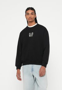 Svart hettegenser laget av bomull med rund hals og ribbestrikkede mansjetter. Har «GAP»-logoen i hvitt på brystet. Båret med lyseblå jeans.