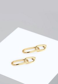 Paire de boucles d'oreilles en chaîne allongée en or reposant sur une surface carrée blanche avec un fond gris.