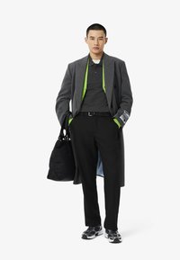 Cappotto grigio con un fodera interna verde, polo nera, pantaloni neri, sneakers nere con dettagli bianchi e una borsa nera. Dettagli in tessuto testurizzato.