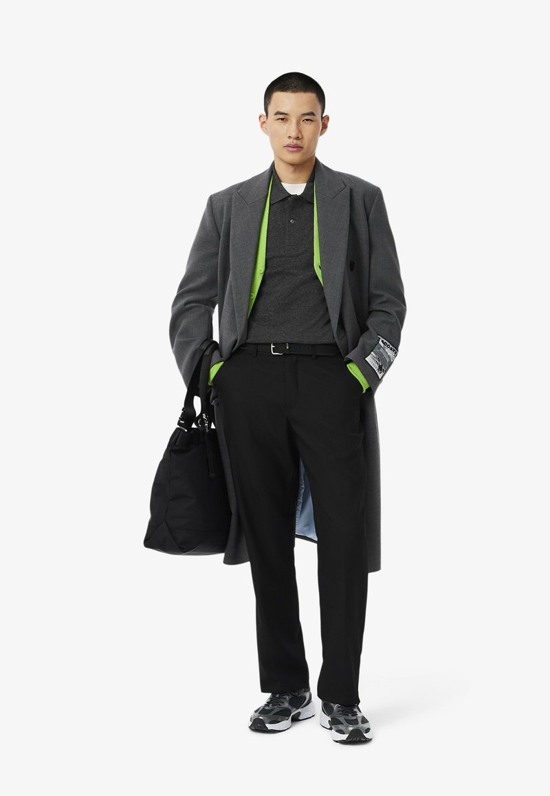 Manteau gris avec une doublure intérieure verte, polo noir, pantalon noir, baskets noires avec des accents blancs, et un sac noir. Détails en tissu texturé.