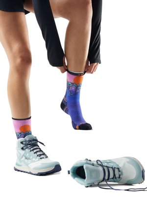 Person wearing colorful mountain-themed socks, putting on one sock, with light blue hiking boots on floor nearby.