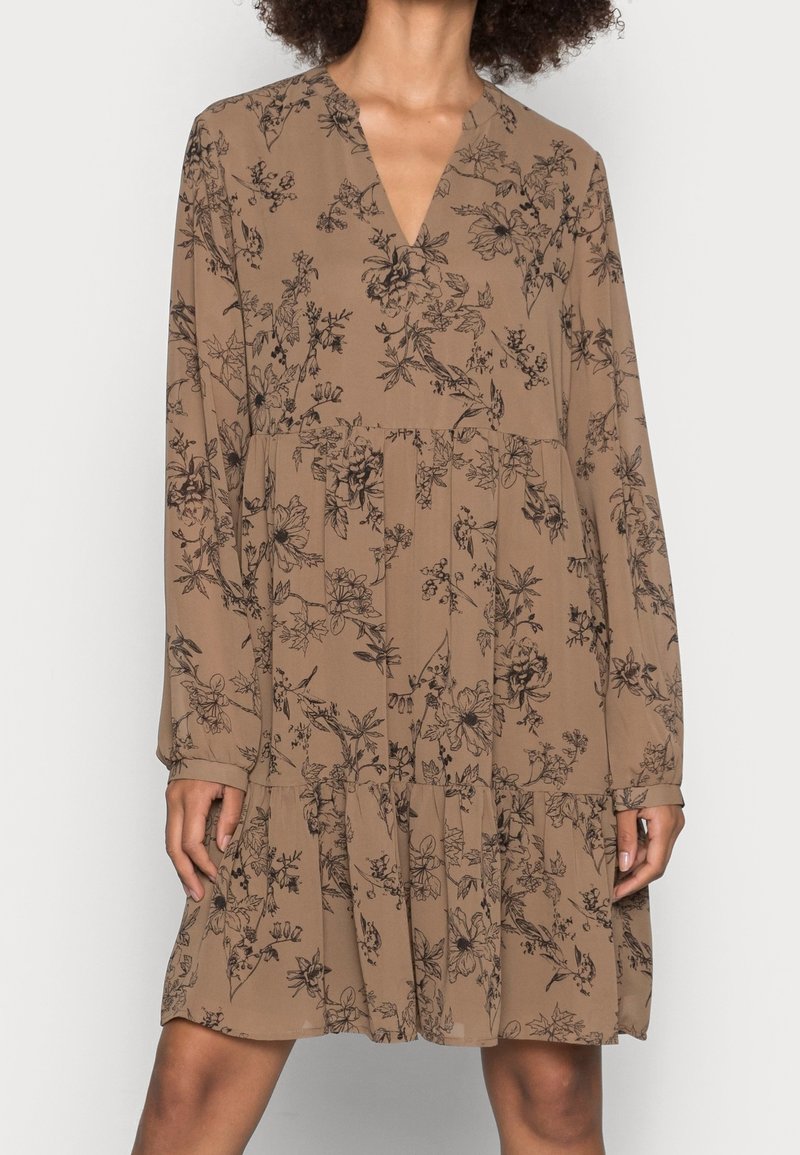 Robe marron à manches longues avec imprimé floral noir, encolure en V et jupe à volants portée par une personne aux cheveux bouclés naturels.
