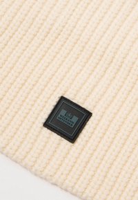 Bonnet tricoté couleur crème avec texture côtelée. Présente un patch carré noir avec l'inscription "WEEKEND OFFENDER" et le logo.