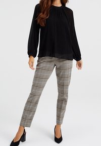 Zwarte geplooide blouse met lange mouwen, gecombineerd met op maat gemaakte geruite broek met beige en bruine patronen, gedragen met zwarte hoge hakken.