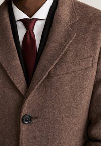 Brązowy płaszcz z mieszanki wełny, z klapami z wycięciem, pojedynczym guzikiem do zapięcia i kieszenią na piersi, w połączeniu z białą koszulą i bordowym krawatem.