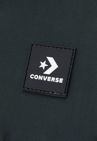 Czarna teksturowana tkanina z prostokątną czarną łatą, na której widnieje białe logo "CONVERSE" oraz wzór gwiazdy i strzały.