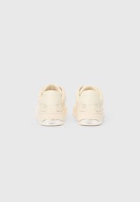 Beige DKNY tossud nähtud tagantpoolt, omades perforeeritud külgdetailide ja hõbedase DKNY brändimisega kanna peal.