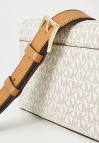 Curea din piele maro cu accesorii aurii atașată la o geantă albă cu un model repetat de logo MK într-o textură subtilă.