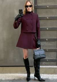 Bordeaux dubbelzijdige jas en rok, kniehoge zwarte stiletto laarzen, zwarte handschoenen, en een kleine zwarte handtas met een klep sluiting.