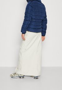 Granatowa puchowa kurtka z poziomym przeszyciem, zestawiona z długą białą spódnicą cargo. Wyróżnia się lekkim materiałem i sportowymi, casualowymi butami.