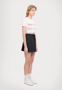 Balts cropped t-krekls ar sarkanu tekstu, apvienots ar melnu plisētu mini svārku. Modelis valkā baltas kedas un gaiši pelēkas zeķes.