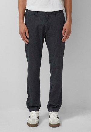 Person trägt dunkelgraue gerade geschnittene Hose und weiße Sneakers mit schwarzen Details, steht vor einem einfarbigen hellgrauen Hintergrund.