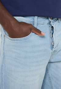 Lichtblauwe denim spijkerbroeken hebben een voorzak waarin een hand rust, wat het klassieke vijf-zakken ontwerp en de knoopsluiting benadrukt.