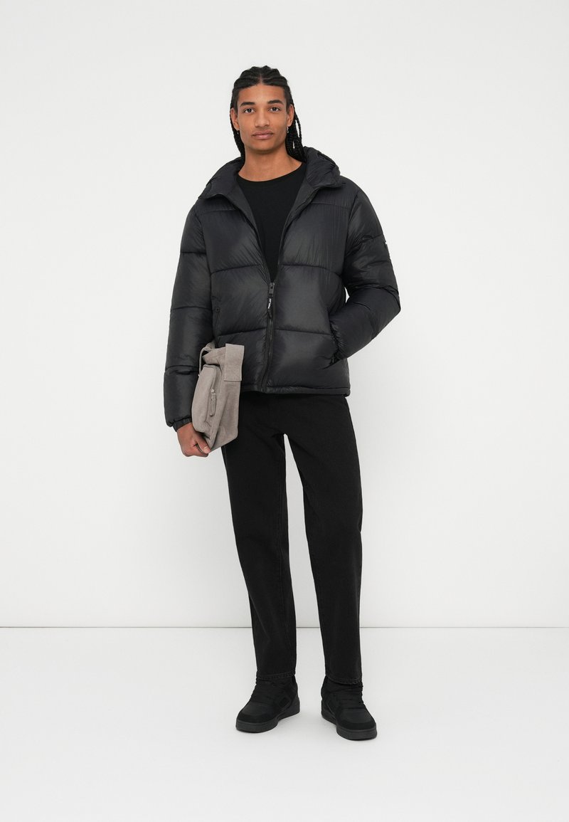 Veste doudoune noire avec capuche, fermeture éclair et design matelassé. Associée à un pantalon noir et des baskets noires, tenant un sac gris.