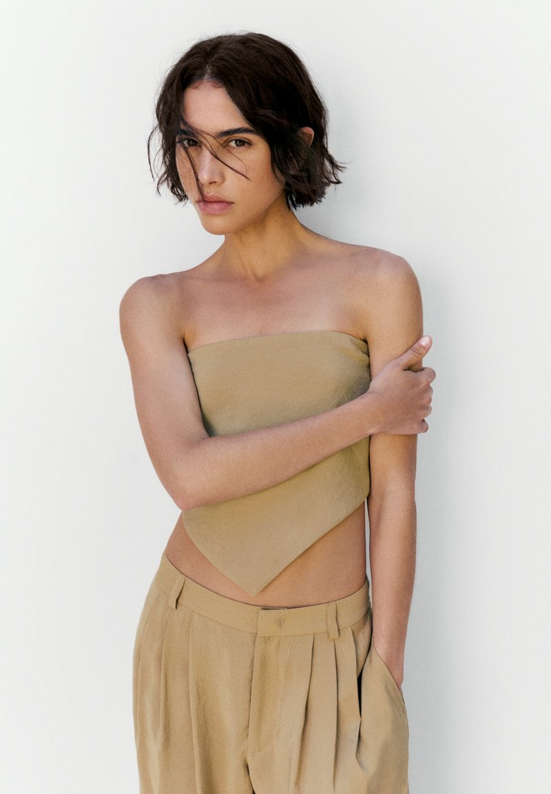 Jeune personne aux cheveux courts et foncés portant un haut beige sans bretelles et un pantalon ample, debout devant un fond blanc uni, le bras croisé.