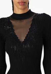 Top lungo a maniche nere a coste con scollo a V in mesh trasparente con bordi scallopati e dettagli in filato metallico. Tessuto testurizzato su tutta la superficie.