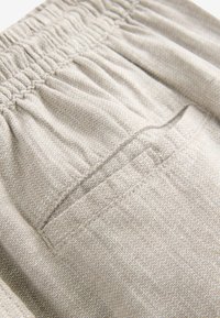 Gros plan sur un tissu beige texturé avec une ceinture élastique et un petit détail de poche cousue.