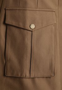 Tissu marron à texture diagonale présentant une grande poche sur la poitrine, fermée par un bouton en métal marqué « Ted Baker ».