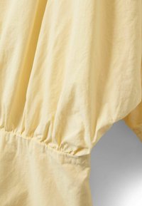 Lysegult stof med en blød tekstur, der har rynkedetaljer ved taljen og flydende ærmer. Glat, rynkebestandigt materiale.
