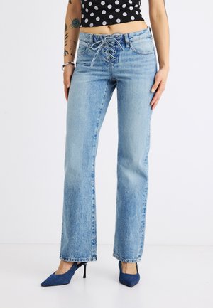 Frau trägt hellblaue Schnür-Jeans, schwarz-weiß gepunktetes Croptop und blaue spitze High Heels, steht vor weißem Hintergrund.