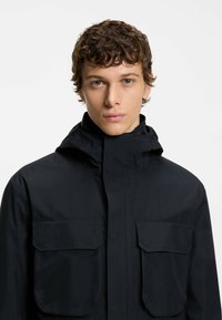Black waterproof jacket with a high collar, two front pockets, and a smooth texture. Features a zip closure and a hood for added protection.