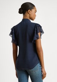 Sötét hajú n�ő kontyba fogott hajjal, tengerészkék pöttyös blúzban fodros rövid ujjakkal és kék farmernadrágban, háttal egy fehér háttér előtt.