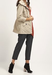 Chaqueta ligera beige con capucha, con detalles de botones, superpuesta a una blusa roja, combinada con pantalones cortos gris oscuro y botas negras de tobillo.