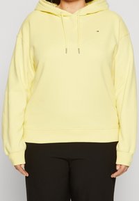 Osoba w pastelowożółtej bluzie z kapturem i czarnych spodniach, stojąca na tle jasnego, jednolitego tła.