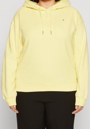 Personne portant un hoodie jaune pastel avec des cordons de serrage et un pantalon noir, se tenant devant un fond clair.