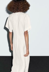 Una persona con capelli ricci è vista di spalle, indossando un abito bianco fluente su uno sfondo minimalista blu chiaro e scuro.