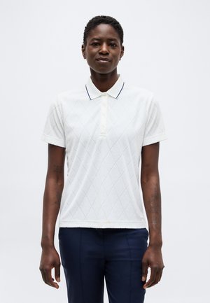 Osoba oblečená v bielej krátkorukávovej polokošeli s diamantovým vzorom a tmavých nohaviciach, stojaca pred jednoduchým svetlým pozadím.