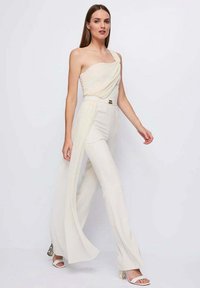 Tuta crema monospalla con corpetto drappeggiato e spacco laterale. Presenta un dettaglio di cintura dorata e tessuto strutturato. Indossata con tacchi bianchi.