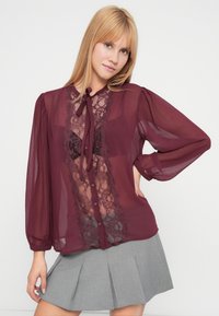 Burgundia värvi läbipaistev pluus pitsdetailidega esiosas, kaelusepaela ja volangiliste varrukatega. Esiosas nähtavad ploomikarva nööbid.