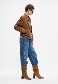 Rjav semiš plašč, ohlapne modre jeans hlače in udobni rjavi čevlji z visokim peto. Model nosi očala in pod njim karirasto srajco.