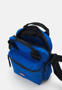 Saco cruzado em tecido azul com múltiplos compartimentos com zíper, alças pretas e um logotipo perto da seção inferior. Material texturizado.