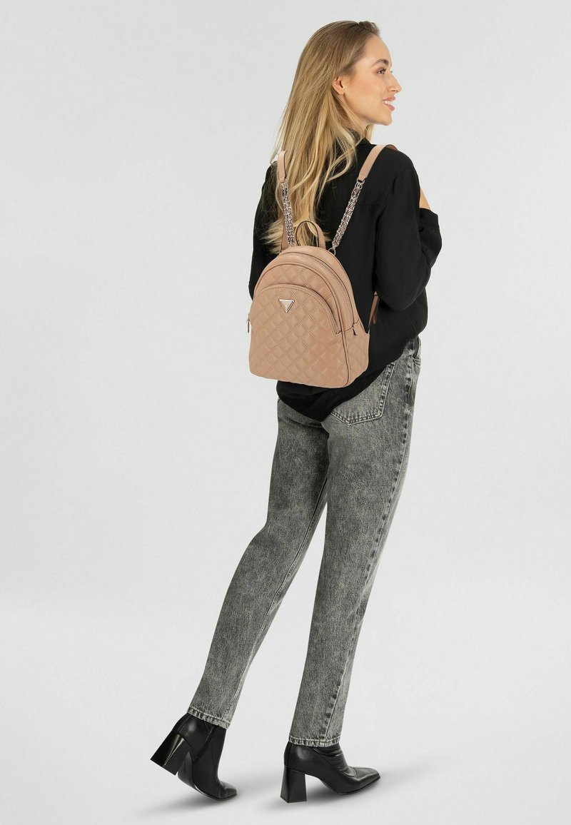 Mochila acolchada color canela con forma redondeada, detalles metálicos y correas de cadena, llevada por una modelo con jeans gris con estampado y zapatos negros.