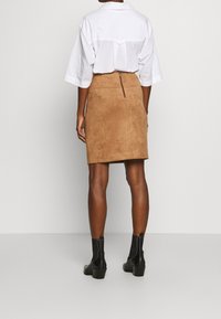 Jupe crayon en daim marron avec une fermeture éclair au dos. Associée à une chemise à manches courtes blanche et des bottines noires. Texture lisse et design ajusté.