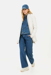 Denimjack met voorzakken en een witte, aansluitende, gevoerde jas. Wijde blauwe denim broek en lichtgekleurde sneakers. Een blauwe pet maakt de look compleet.