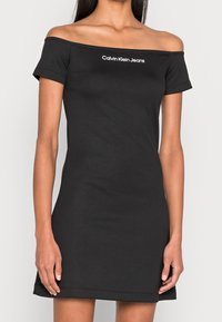 Kvinna i en figurnära svart off-shoulder klänning med korta ärmar och "Calvin Klein Jeans" loggan centrerad på bröstet.