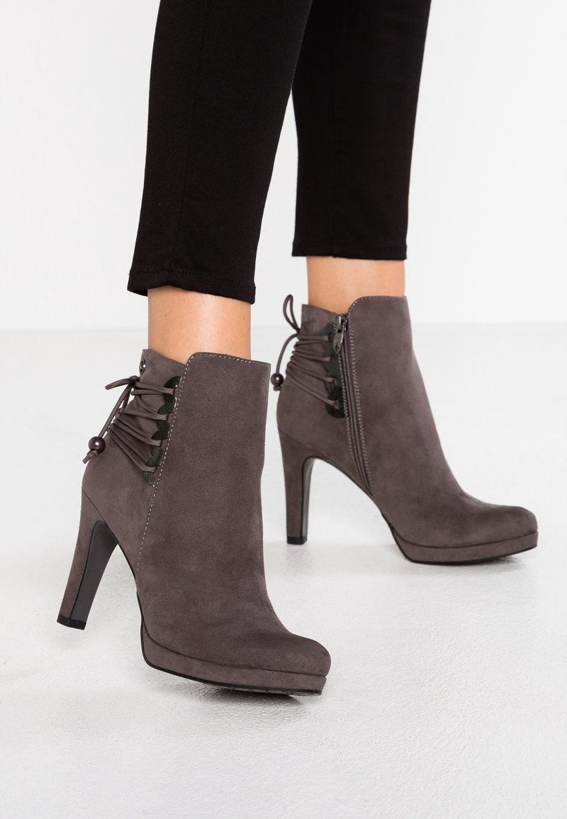 Bottines à talons hauts en daim gris avec fermeture éclair latérale et lacets décoratifs, portées avec un pantalon noir court sur un sol blanc.